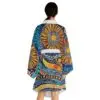 A woman with dark hair in a low ponytail wears the Vibrant Long Sleeve Kimono Robe, featuring bold abstract sunburst and geometric patterns in orange, blue, yellow, and black; she stands with her back to the camera.